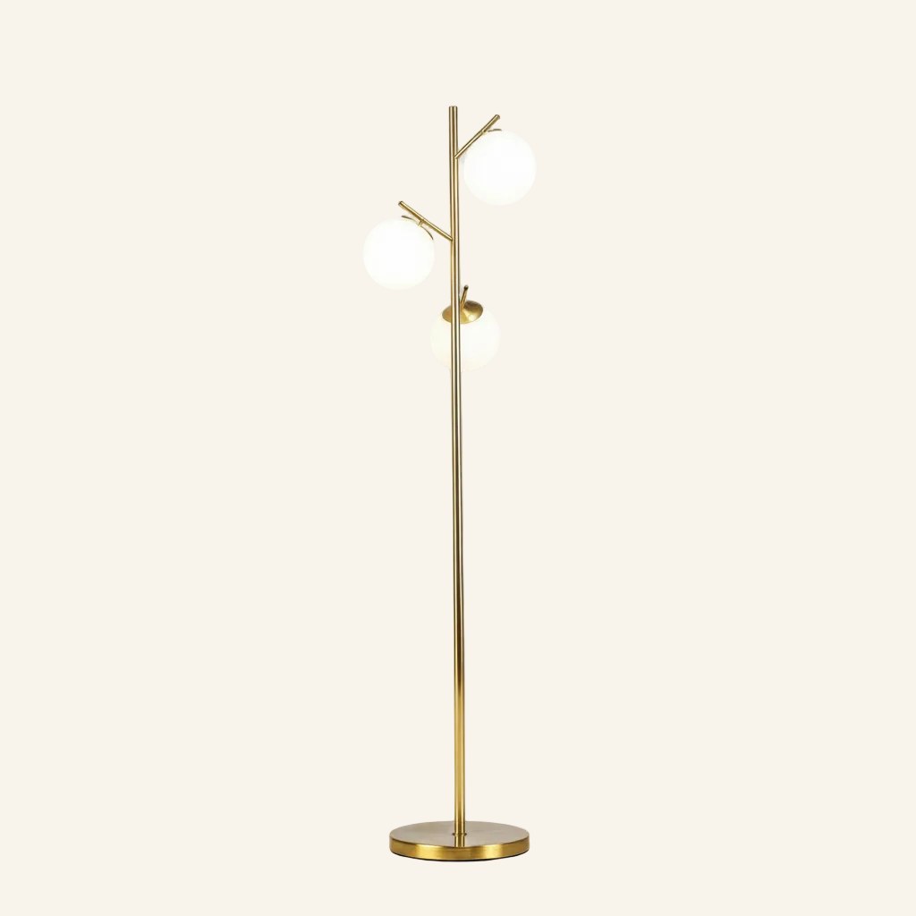 Slim brushed brass floor lamp with three opal white glass globes on angled branch arms, rising from a round metal base on cream backdrop.