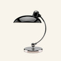 Glossy black domed shade cantilevered from a curved chrome arm on a round polished base, Bauhaus table lamp shown on cream backdrop.