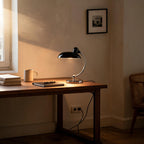 Lit black dome lamp on a wooden desk beside a stoneware mug, notebook and pen, with framed art and caned chair in a sunlit bedroom corner.