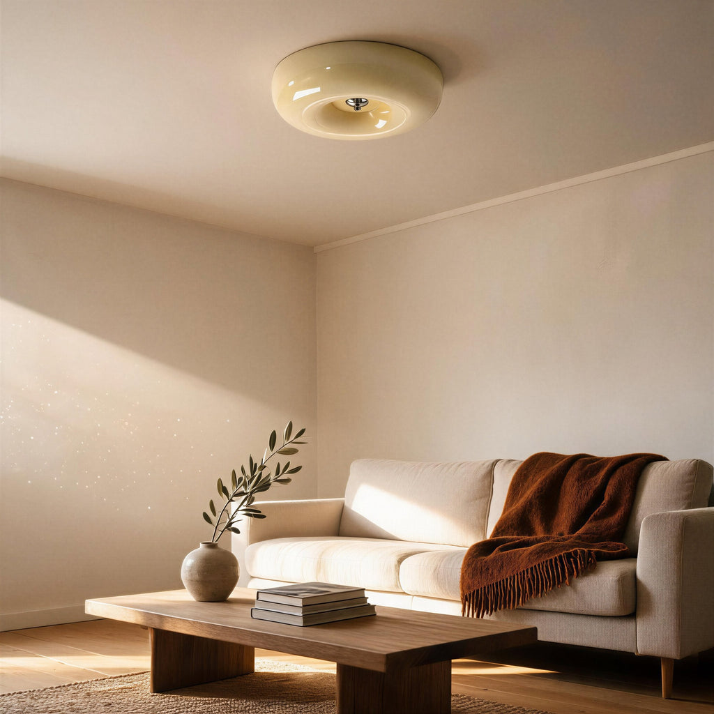 Cream opal flush mount glowing on a neutral living room ceiling above a linen sofa, rust throw, wooden coffee table and olive sprig.