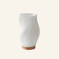 Curvy white 3D-printed table lamp with twisted ribbed shade on a round turned wood base, shown on cream studio background.