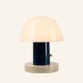 Opal glass dome shade glowing warmly above a navy cylindrical stem and cream disc base, brass touch button on the side