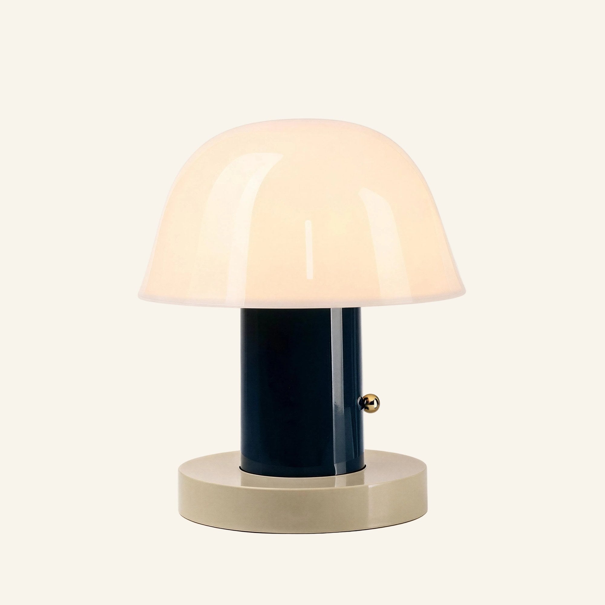 Opal glass dome shade glowing warmly above a navy cylindrical stem and cream disc base, brass touch button on the side