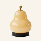 Pear-shaped cream glass table lamp with brass touch-dimmer finial and black cylindrical base, shown on a soft cream studio backdrop