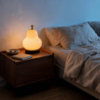 Lit pear glass lamp glowing warm on a walnut nightstand beside linen bedding, casting cozy bedside light in a japandi bedroom
