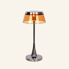 Lamp switched on with warm amber glow filling the tinted acrylic shade, highlighting the chrome stem and weighted disc base