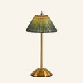 Cordless table lamp with pleated green ribbed-glass shade, brass stem and dome base, illuminated against a cream studio backdrop.