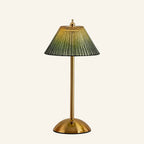 Cordless table lamp with pleated green ribbed-glass shade, brass stem and dome base, illuminated against a cream studio backdrop.