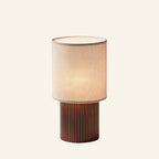 Cordless table lamp with cream linen cylinder shade and fluted copper-brown aluminum base, shown on cream studio background.
