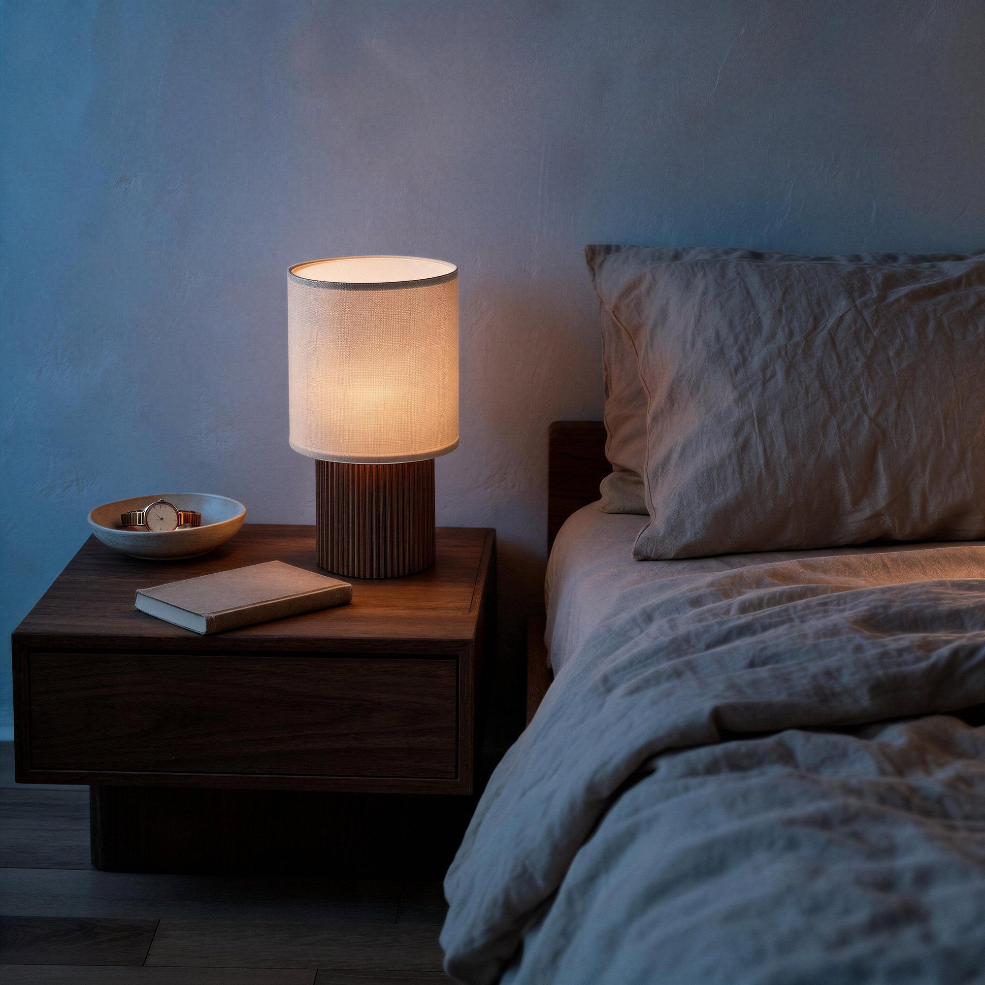 Lit portable lamp on walnut nightstand beside linen-dressed bed, casting warm light against moody blue bedroom wall at dusk.