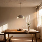 Dome pendant lamp glowing softly above an oak dining table with woven mats, fig bowl, wine glass and stoneware vase in sunlit dining room.