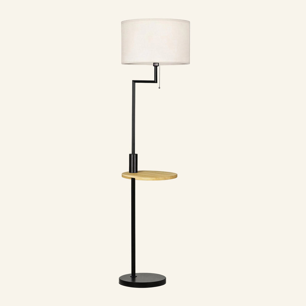 Matte black metal floor lamp with white linen drum shade, integrated round bamboo shelf and pull-chain switch on cream background.