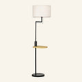Matte black metal floor lamp with white linen drum shade, integrated round bamboo shelf and pull-chain switch on cream background.