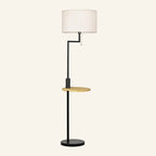 Matte black metal floor lamp with white linen drum shade, integrated round bamboo shelf and pull-chain switch on cream background.