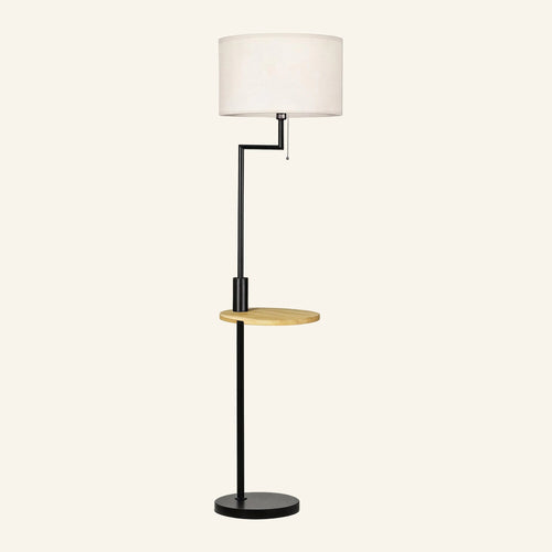 Matte black metal floor lamp with white linen drum shade, integrated round bamboo shelf and pull-chain switch on cream background.