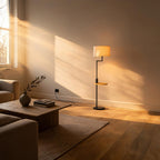 Lit floor lamp with bamboo side shelf glowing softly beside a low wood coffee table in a sunlit minimalist living room.