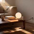 Lit bedside table lamp on a walnut coffee table beside stacked books and a small vase in a sunlit living room.