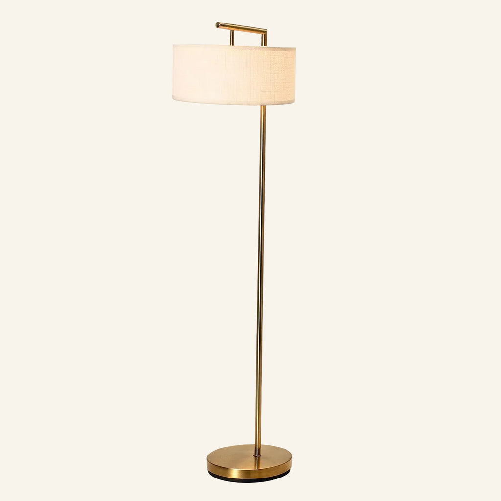 Slim brass floor lamp with cantilevered arm holding a white linen drum shade above a round weighted base on cream backdrop.
