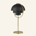 Matte black twin-dome shades rotate within a brass ring on a slim brass stalk and circular base, sculptural table lamp on neutral background.