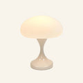 Cordless mushroom table lamp glowing warmly, frosted white dome shade above a glossy ivory tulip base on cream backdrop