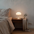 Lit mushroom lamp on a wooden nightstand beside linen bedding, olive sprig and watch tray creating a calm minimalist bedroom scene