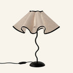 Scalloped linen petal shade with black trim sits atop a wavy black iron stem and domed base, studio shot on cream background.
