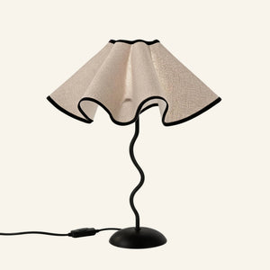 Scalloped linen petal shade with black trim sits atop a wavy black iron stem and domed base, studio shot on cream background.