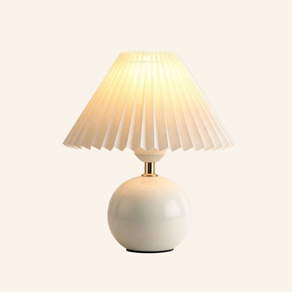 Illuminated table lamp with white pleated fabric shade, polished brass neck, and glossy spherical ceramic base on cream backdrop