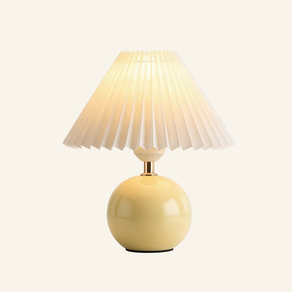 Soft butter-yellow ceramic sphere base supporting a fan-pleated ivory shade, compact bedside lamp shown on neutral background