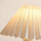 Close-up of the crisp accordion pleats on the ivory fabric shade, glowing warmly to reveal woven texture and brass-trimmed neck