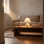 Pleated ceramic table lamp glowing on an oak coffee table beside an olive sprig and stacked books in a sunlit linen-toned living room
