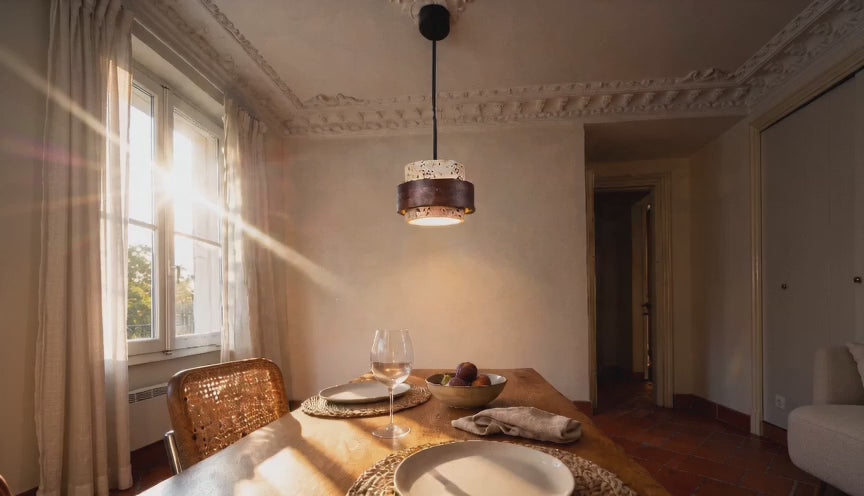 Cinematic video of a two-tier travertine and walnut wood pendant lamp glowing warmly above a sunlit dining table.