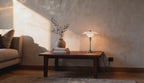 Cinematic video of a tiered opal glass table lamp glowing softly on a walnut coffee table beside an olive branch in a sunlit living room.