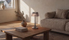 Cinematic video of a cordless chrome table lamp with tinted acrylic drum shade glowing on a wooden coffee table beside a sofa.
