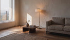 Cinematic video of a gold steel floor lamp with white linen drum shade glowing in a sunlit living room beside a low wooden coffee table.