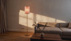 Cinematic video of a slim cream linen drum floor lamp glowing beside a textured sofa in a warm living room at dusk.