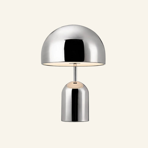 Polished chrome mushroom-shaped table lamp with domed shade and cylindrical base, glowing warmly on a cream studio backdrop