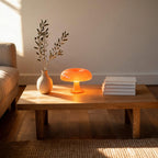 Lit orange mushroom table lamp on an oak coffee table beside a stoneware vase of olive branches and a stack of books in a sunlit living room.