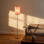 Lit floor lamp casting warm glow in a living room beside a linen sofa, walnut coffee table, ceramic vase with olive branch, and woven rug.