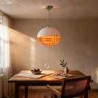 Pendant lamp glowing warm amber above a walnut dining table set with figs, wine glass and olive sprig, cane chair and linen curtains nearby.