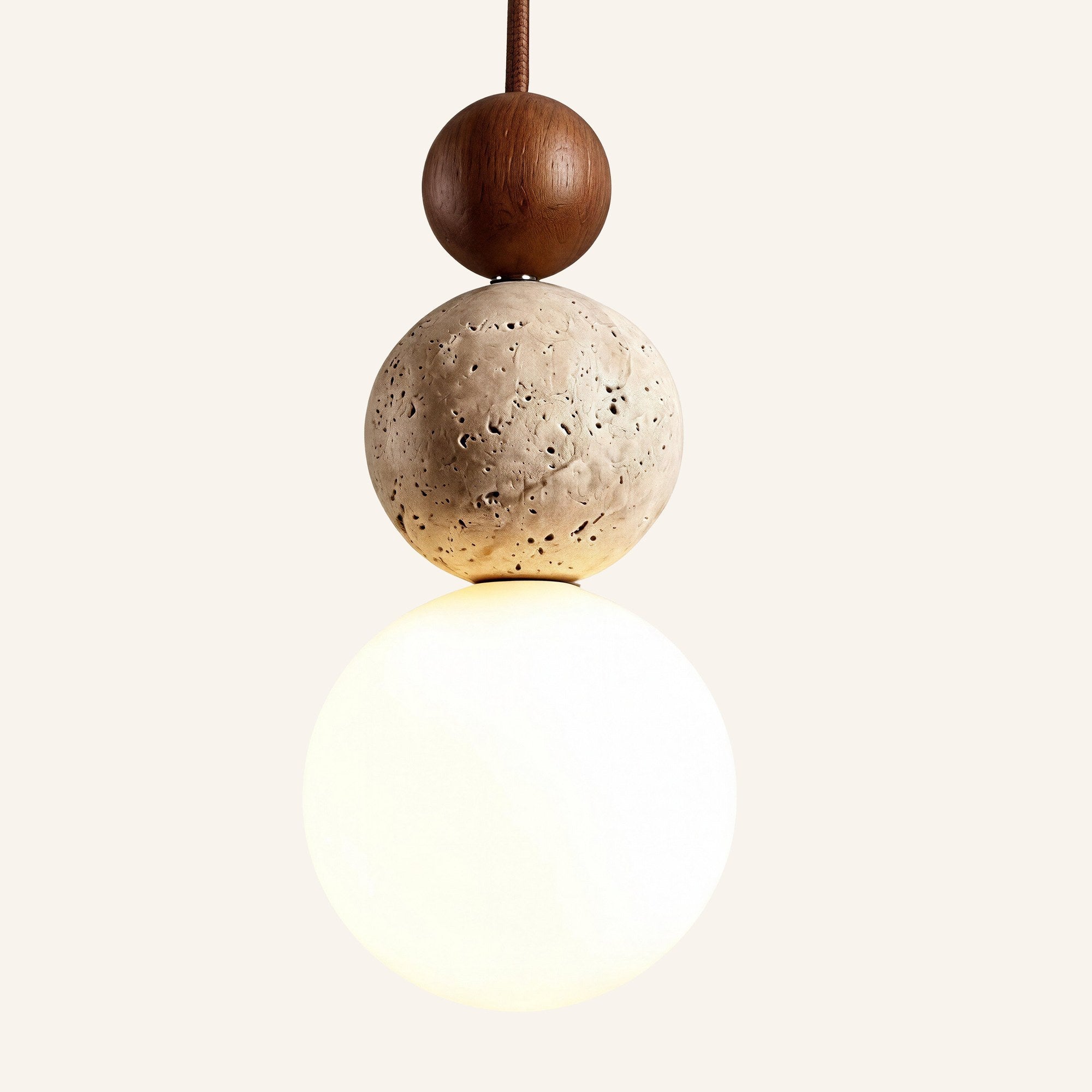 Stacked pendant lamp with walnut sphere, pitted travertine stone orb and frosted opal glass globe glowing warm on cream backdrop.