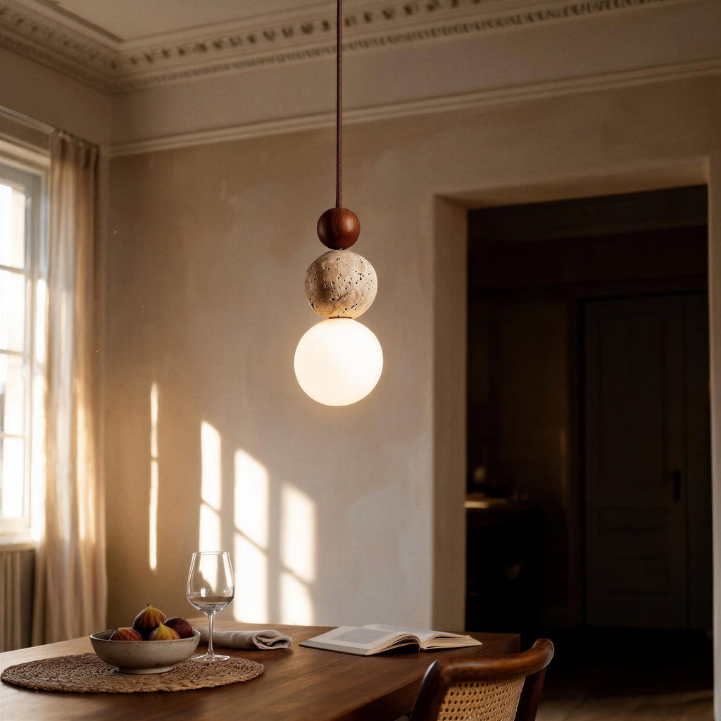 Pendant lamp hanging above wooden dining table with fig bowl, wine glass and open book in sunlit plaster-walled bedroom corner.