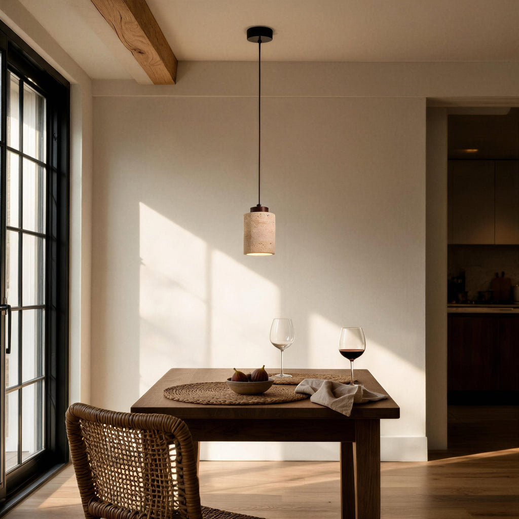 Travertine pendant glows above a wooden dining table set with wine glasses and figs, sunlit room with beamed ceiling and rattan chair.