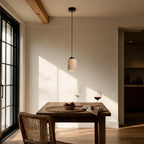 Travertine pendant glows above a wooden dining table set with wine glasses and figs, sunlit room with beamed ceiling and rattan chair.