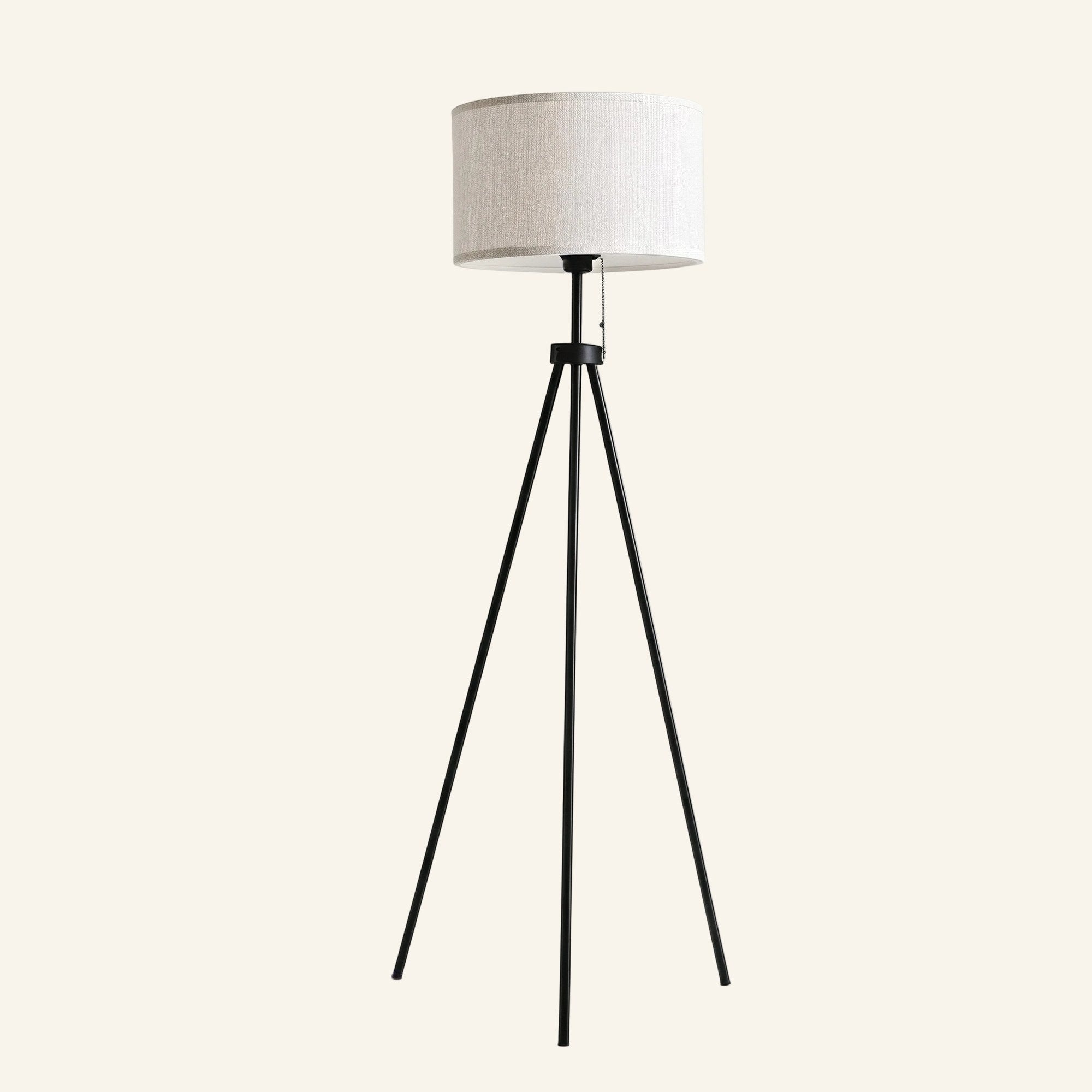 Tripod floor lamp with white linen drum shade on three slim black metal legs, with pull-chain cord, on cream background.