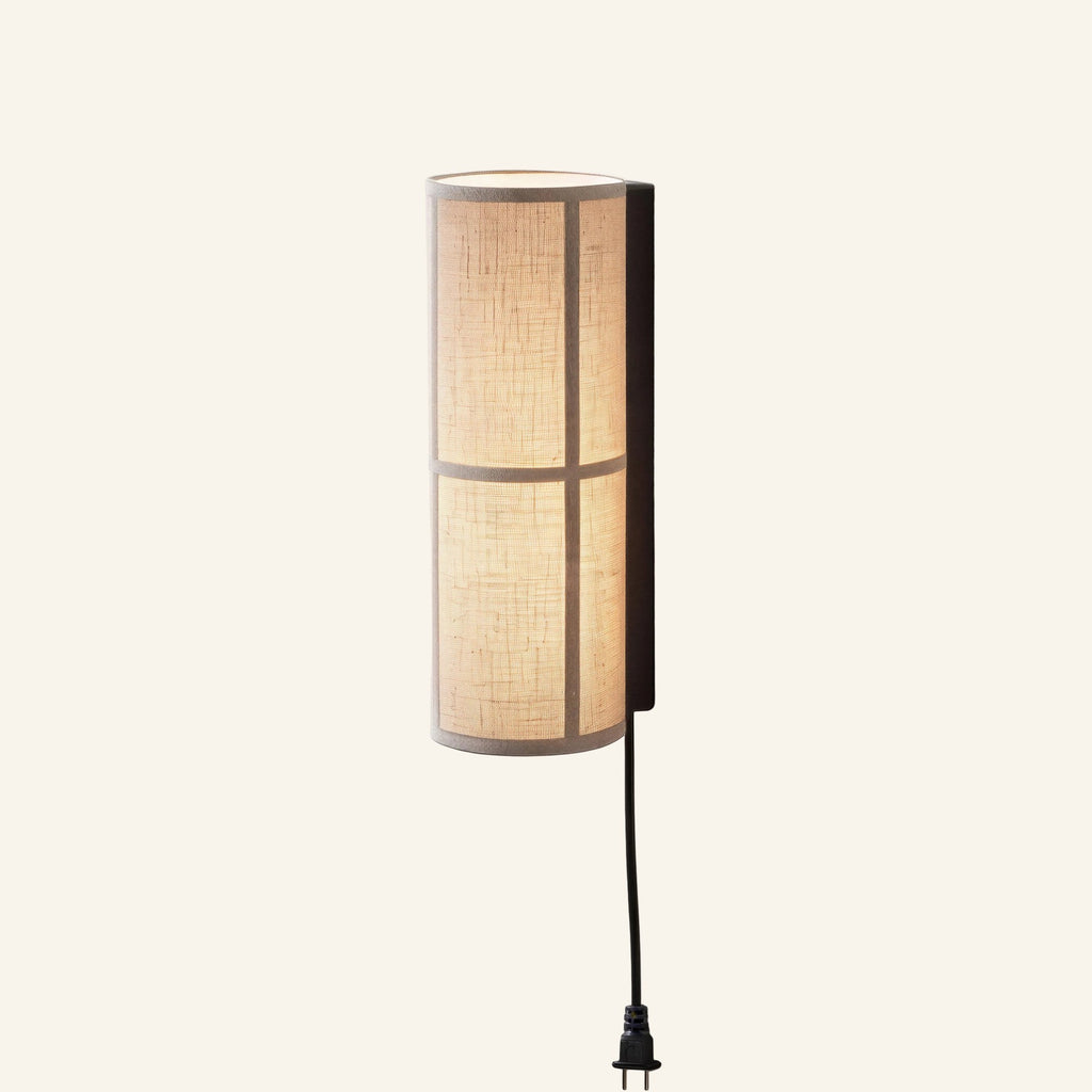 Cylindrical beige linen wall sconce on a black bracket with plug-in cord, glowing warmly against a cream background.