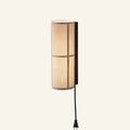 Cylindrical beige linen wall sconce on a black bracket with plug-in cord, glowing warmly against a cream background.