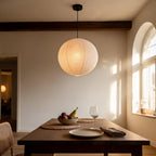 Rice paper globe pendant lamp hanging over a wooden dining table at sunset, with fruit bowl and wine glass below.